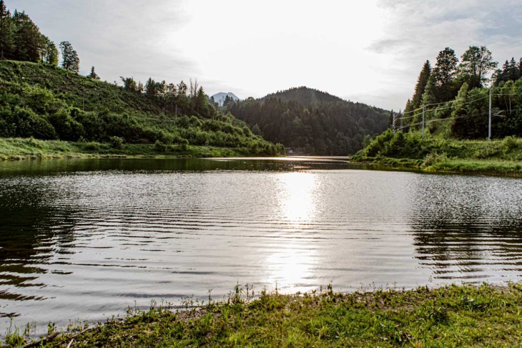Ein ruhiger See, umgeben von grünen Hügeln und Bäumen, mit der Sonne, die hell über die fernen Berge scheint und sich auf der Wasseroberfläche spiegelt.