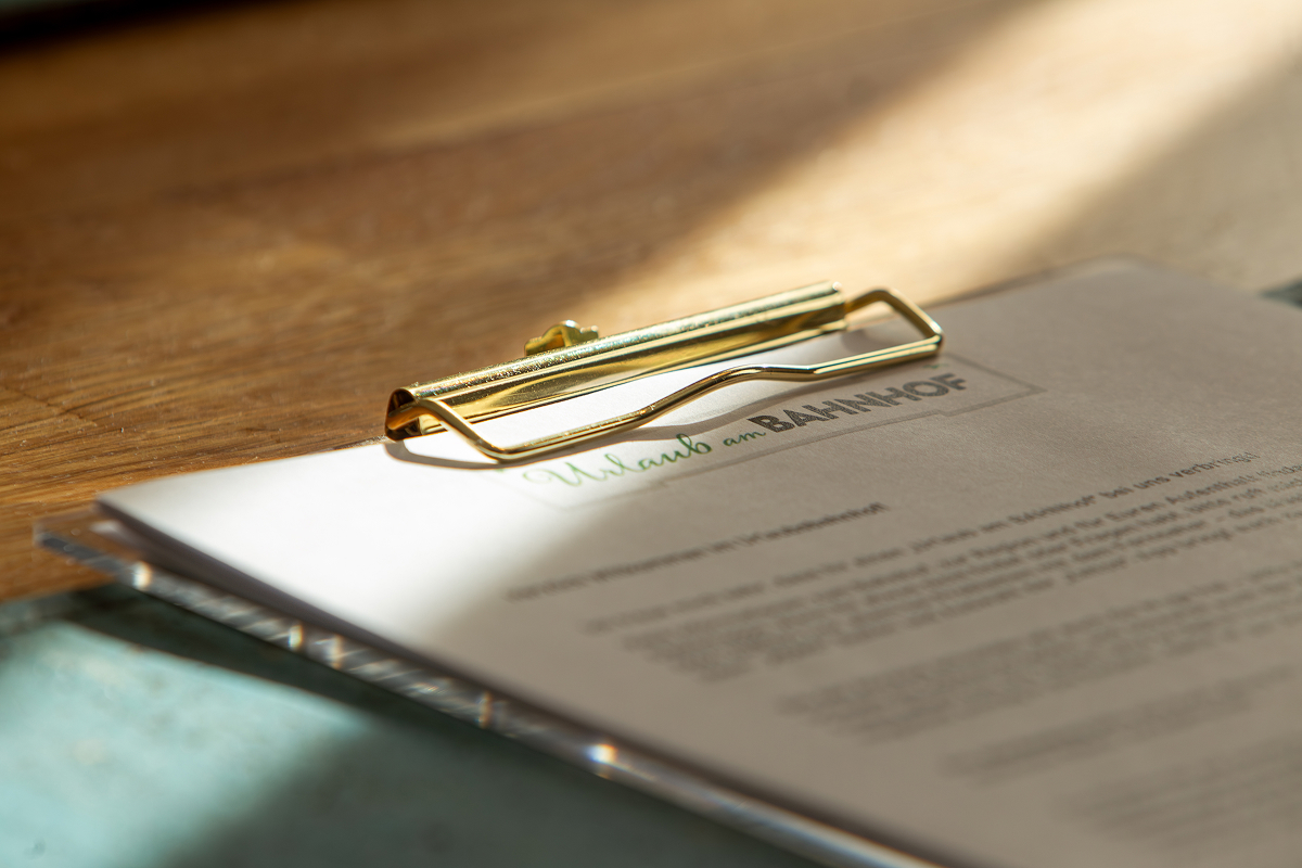 Nahaufnahme eines Klemmbretts, auf dem ein Dokument mit der Überschrift Urlaub am BAHNHOF auf einer hölzernen Oberfläche liegt, wobei das Sonnenlicht Schatten auf das Papier wirft.