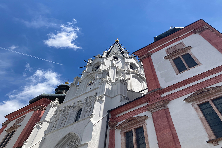 Eine hohe, verschnörkelte Kirche mit weißen und rosaroten Verzierungen steht vor einem strahlend blauen Himmel mit Wolkenfetzen, gesehen aus einem niedrigen Winkel. Ein Flugzeug hinterlässt einen Kondensstreifen am Himmel über der Kirchturmspitze.