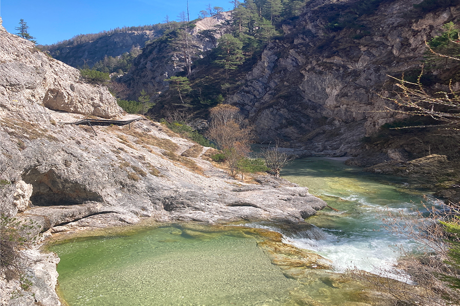 Ein klarer Bach mit kleinen Wasserfällen fließt durch eine felsige Bergschlucht, umgeben von steilen Felsen und grünen Bäumen unter einem strahlend blauen Himmel.