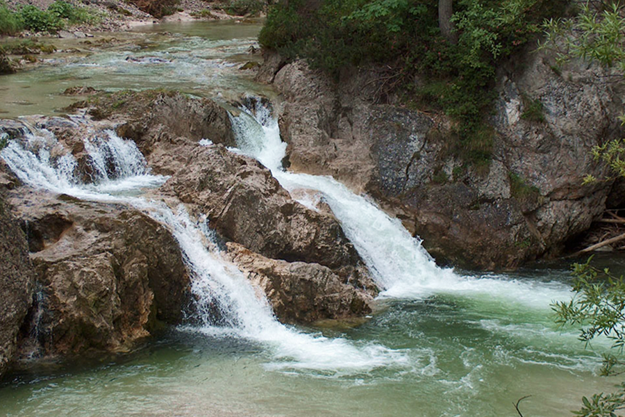 Kleine Wasserfälle stürzen über Felsformationen in einen grünlichen Fluss, der von üppigen Bäumen und Pflanzen umgeben ist. Die Szene wirkt natürlich und ruhig, mit fließendem Wasser und zerklüfteten Felsen.